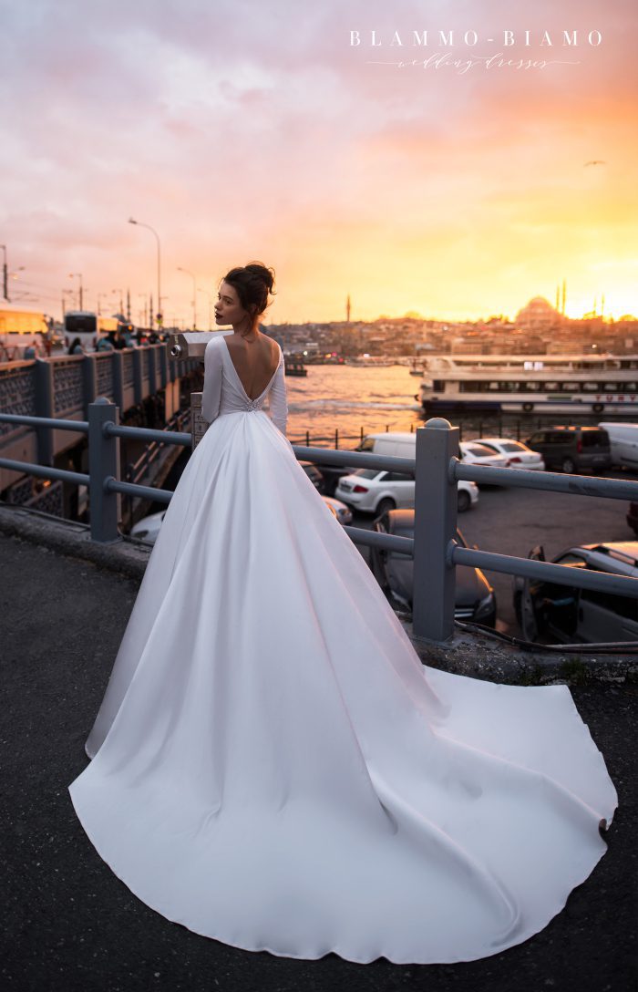 A bride wearing satin princess wedding dress Tilda by blammo-biamo with long sleeves, long train and open back with brooch by Rara Avis at Dell' Amore Bridal, Auckland, NZ. 2